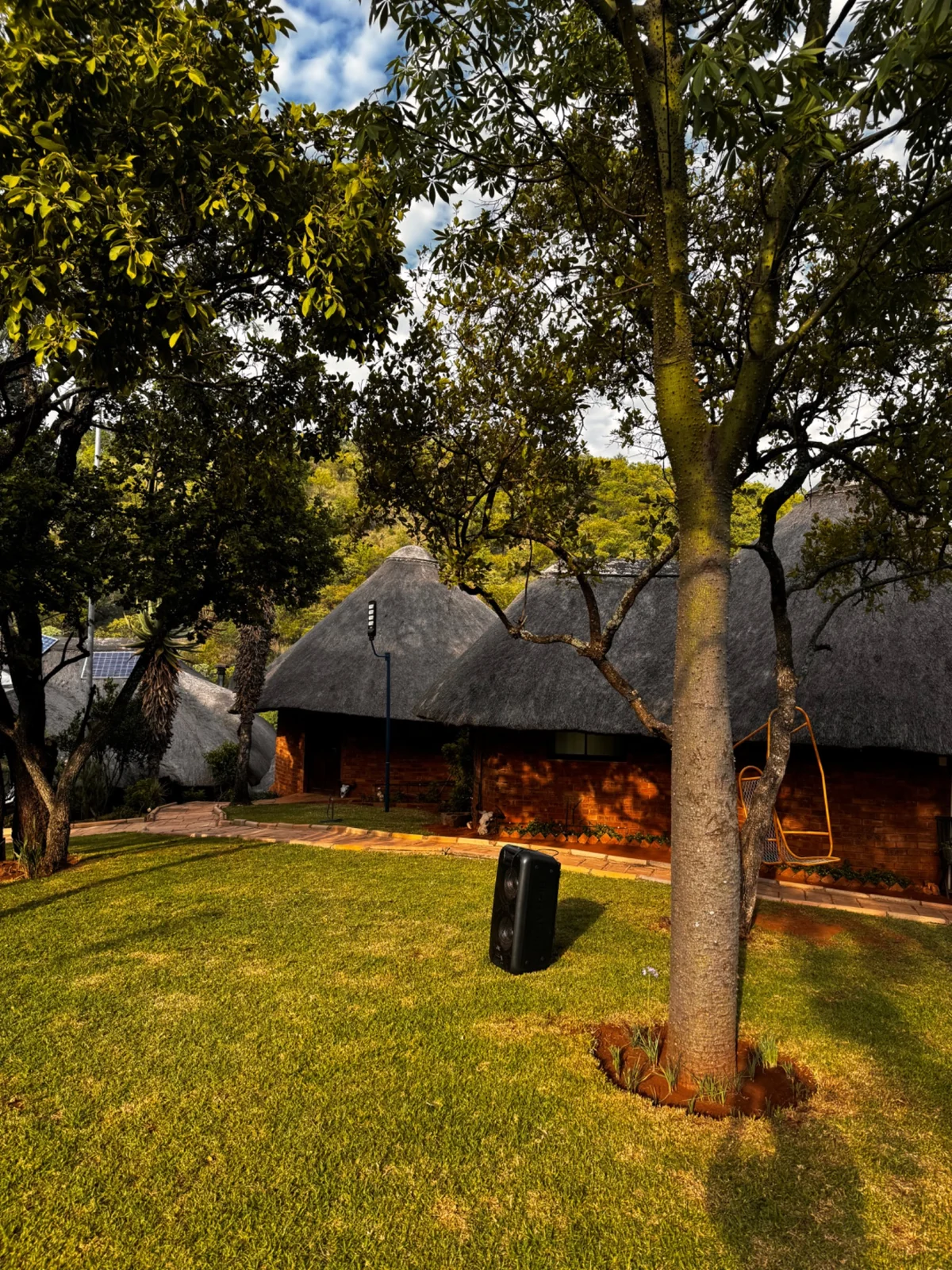 Thatched lodge buildings in a green courtyard beneath tall trees.